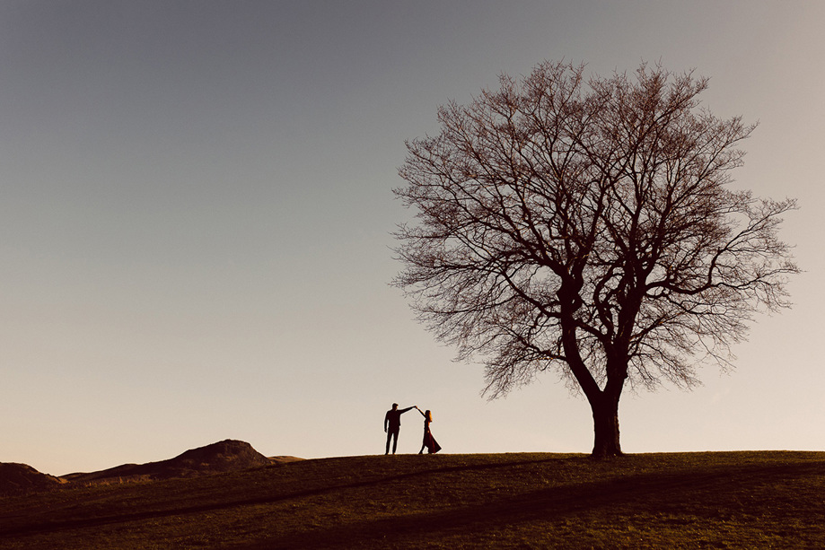 Groom spins his bride at sunset on a hill with a large bare tree beside them