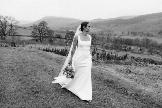 Bride walking across grass with bouquet in hand and hills in background