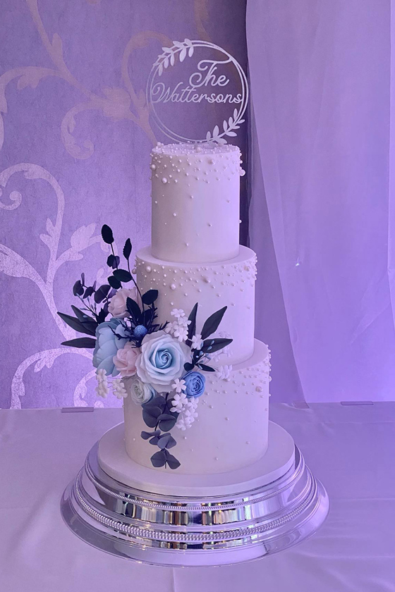 White three-tier cake decorated with pearls and blue pink and white flowers with cake topper that reads "The Wattersons"