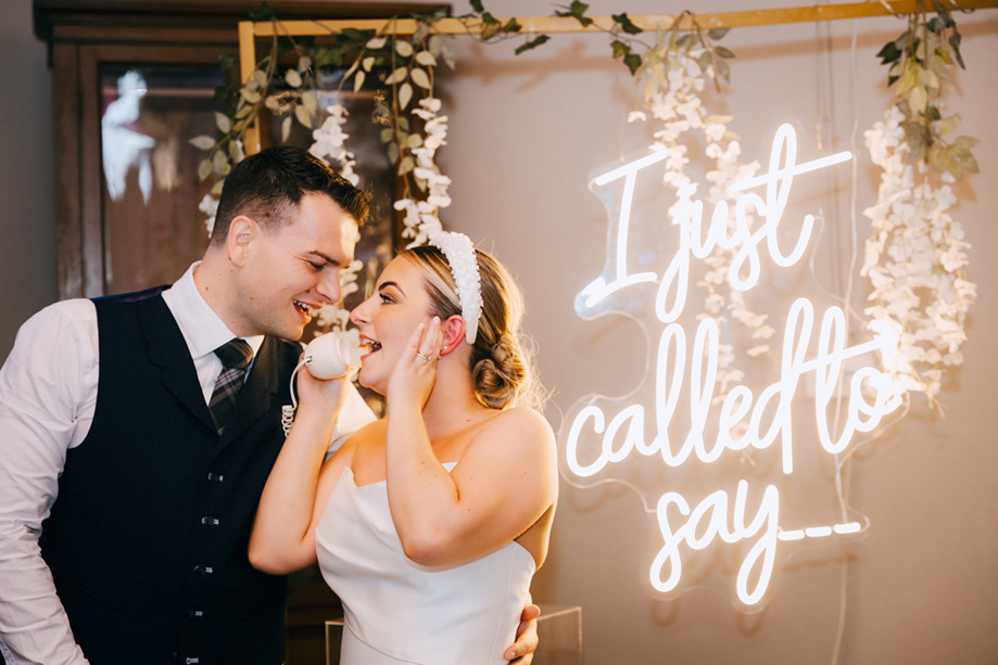 Bride and groom laugh as they leave a message on their audio guest book with "I just called to say..." sign in background