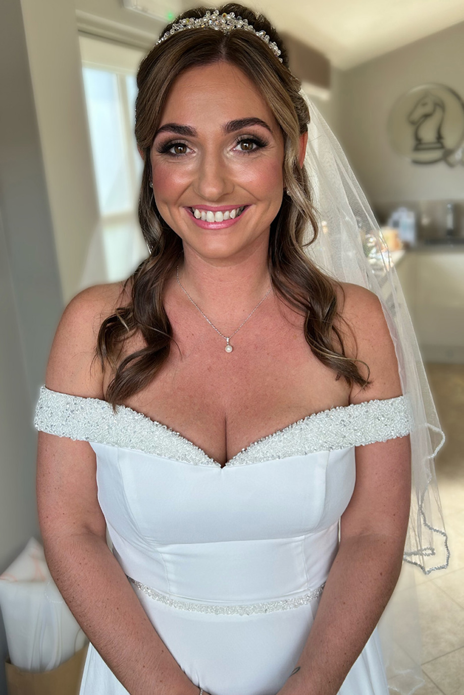 Bride with bronzed skin and baby pink lipstick smiles in her dress and veil with pearl tiara