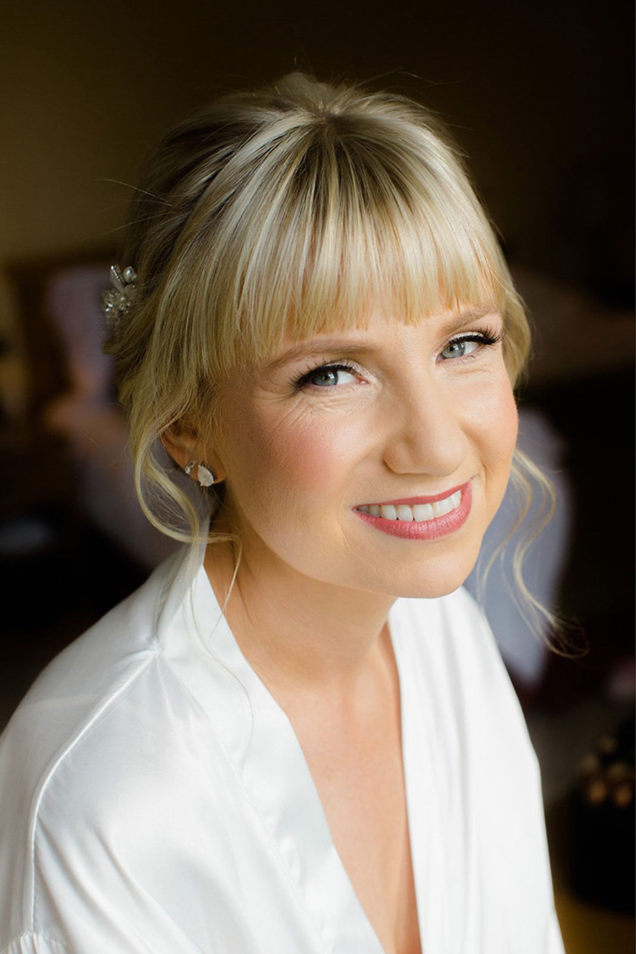 Blonde bride with fringe smiles at the camera with coral lipstick and blush