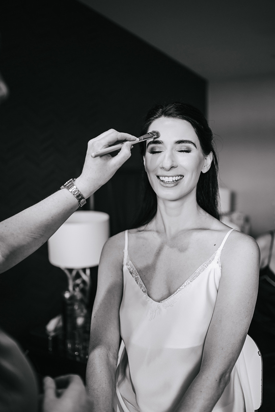 Black and white image of bride's makeup being applied and smiling with her eyes closed