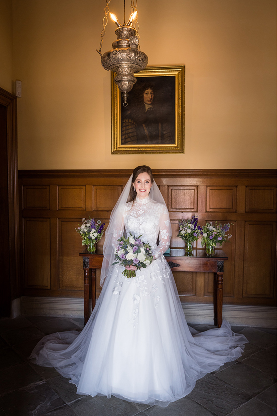 Bride smiles and stands in front of painting in her lace dress and veil holding her bouquet