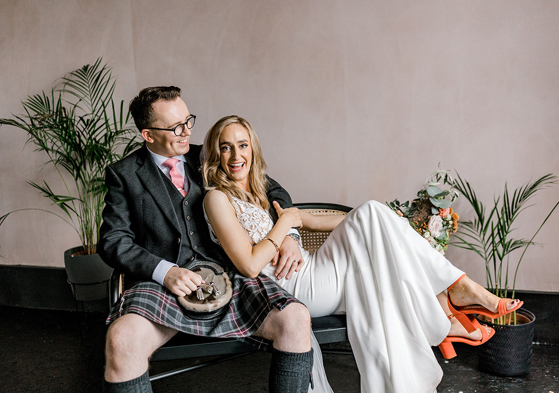 Bride and groom laughing together on a rattan bench, with the groom wearing a kilt and the bride in a white wedding outfit with orange heels.