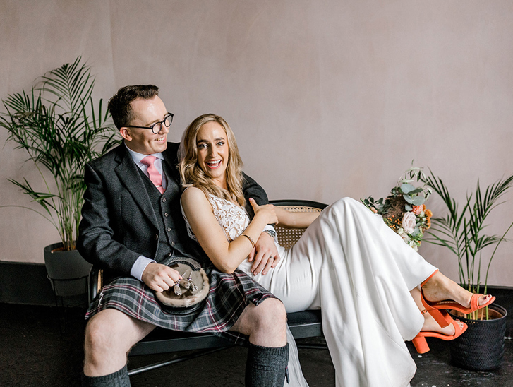 Bride and groom laughing together on a rattan bench, with the groom wearing a kilt and the bride in a white wedding outfit with orange heels.