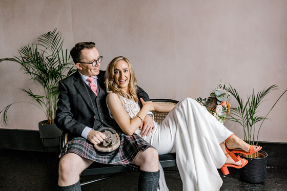 Bride and groom laughing together on a rattan bench, with the groom wearing a kilt and the bride in a white wedding outfit with orange heels.