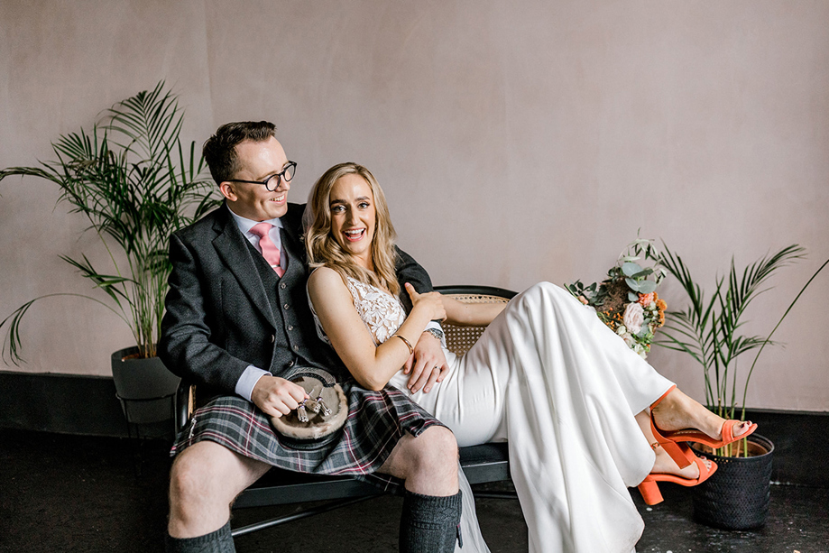 Bride and groom laughing together on a rattan bench, with the groom wearing a kilt and the bride in a white wedding outfit with orange heels.
