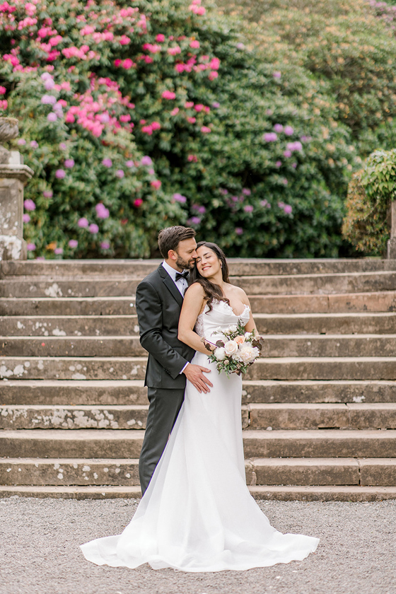 Bride and groom in front of stone steps with pink garden flowers at Auchen Castle wedding venue