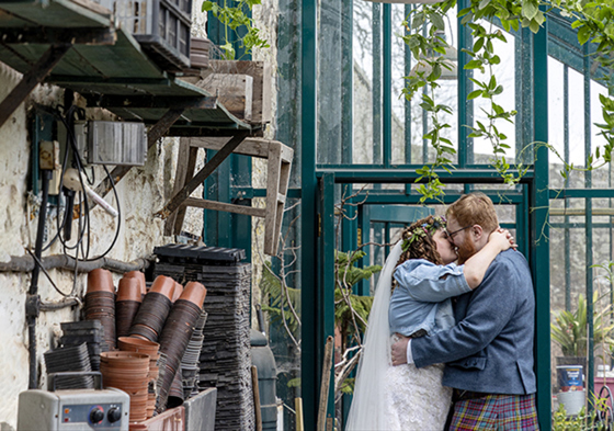 Bride in flower crown and denim jacket kisses her groom in a kilt inside greenhouse-style room