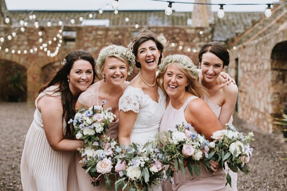 Bridal party laugh together in courtyard of wedding venue, two are wearing flower crowns and they are all holding bouquets and wearing pink dresses. Makeup is by Jennifer Louise Studio.