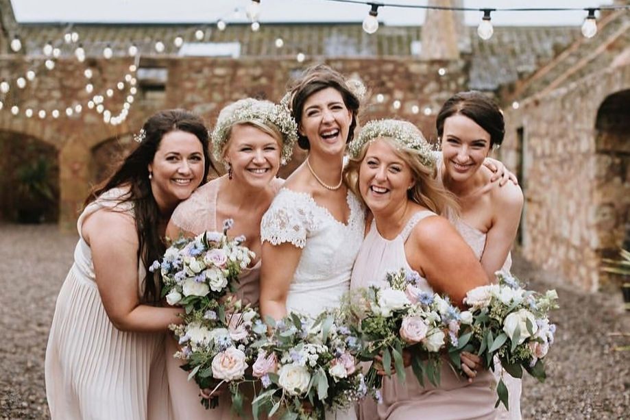 Bridal party laugh together in courtyard of wedding venue, two are wearing flower crowns and they are all holding bouquets and wearing pink dresses. Makeup is by Jennifer Louise Studio.