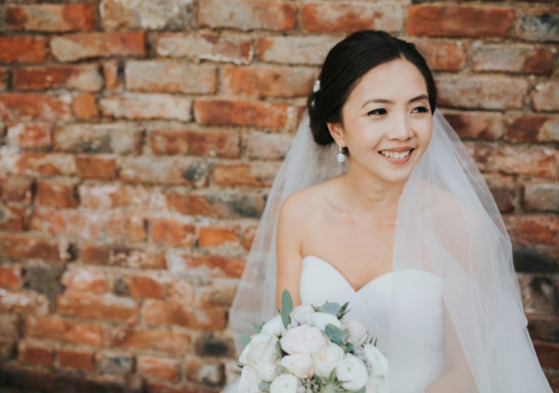 Bride sits in front of a brick wall and smiles off to the side, makeup by Jennifer Louise Studio.