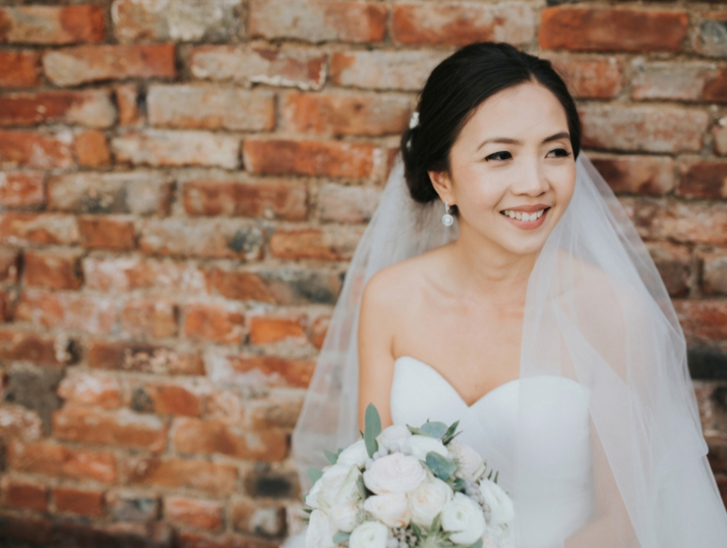 Bride sits in front of a brick wall and smiles off to the side, makeup by Jennifer Louise Studio.