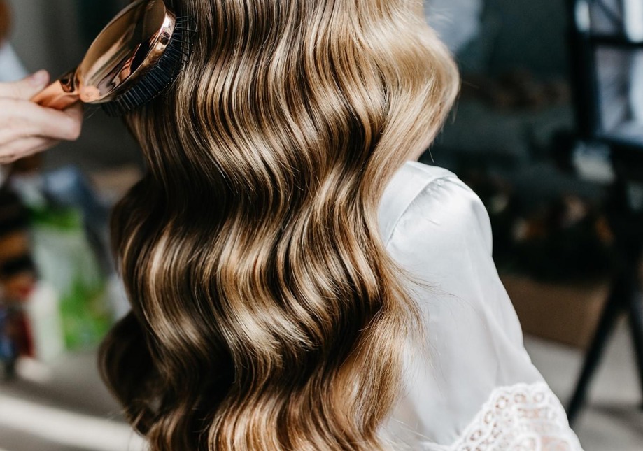 A bride is wearing a silk and lace dressing gown, having her hair styled in soft waves, radiating joy and anticipation for her upcoming wedding day.