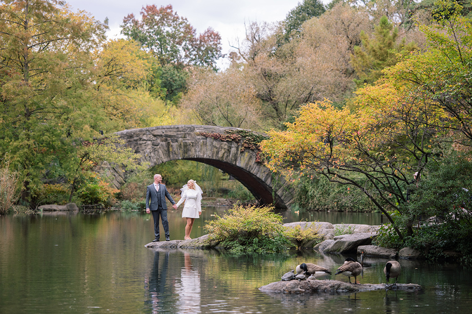 Bride and groom standing by the water in Central Park with historic stone bridge behind them