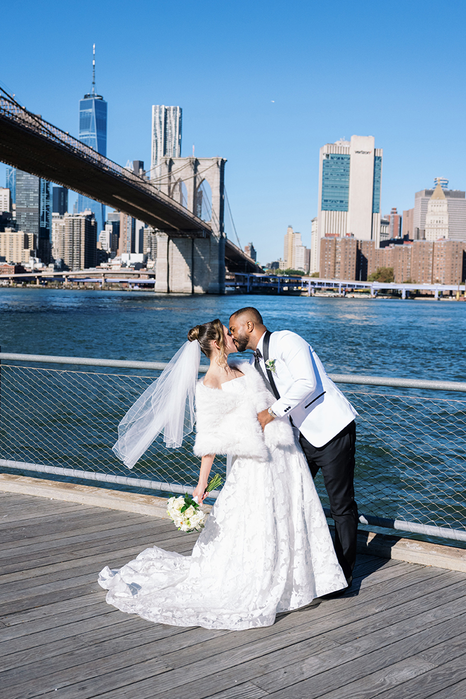 Groom dipping bride for a kiss by the water with Brooklyn Bridge and NYC skyline behind