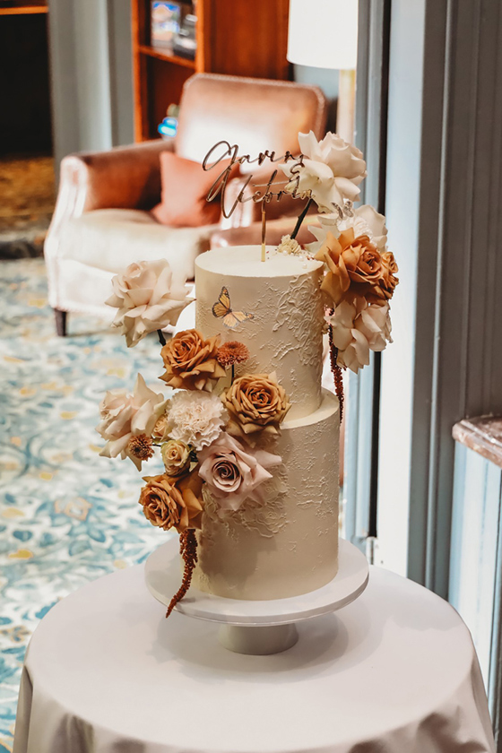 Two tier cream cake with butterfly details and pastel and rust-coloured roses with black cake topper by YiShi Cake