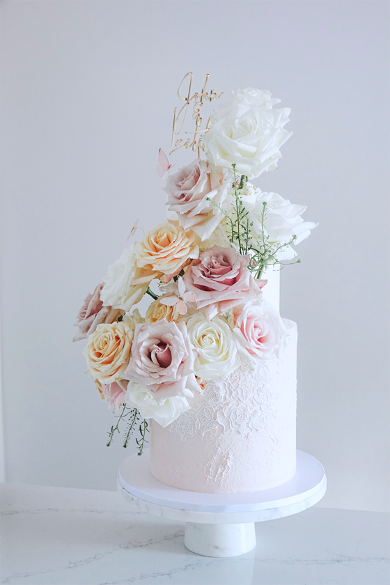 Baby pink two-tier cake with lace detailing, pastel flowers and gold cake topper by YiShi Cake