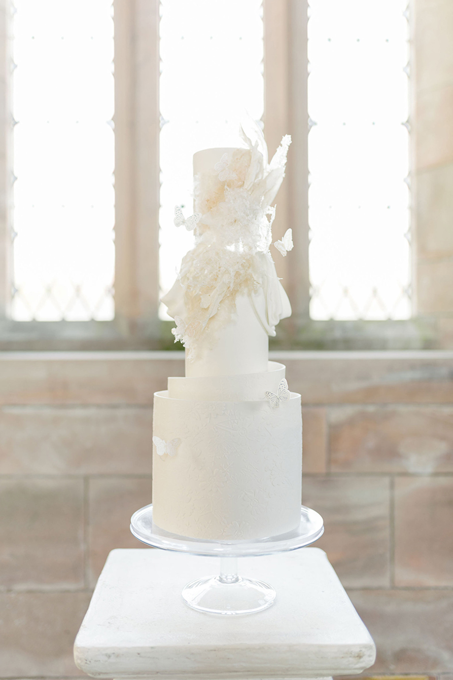 White three-tier cake on transparent stand with signature ruffles and butterfly detailing by YiShi Cake