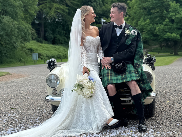 Bride and groom looking at each other sit on bonnet of a cream wedding car with black bows, captured by Scottish wedding content creator The Perfect Day Club.
