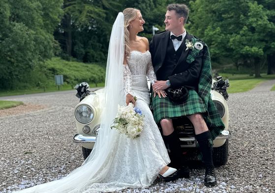 Bride and groom looking at each other sit on bonnet of a cream wedding car with black bows, captured by Scottish wedding content creator The Perfect Day Club.