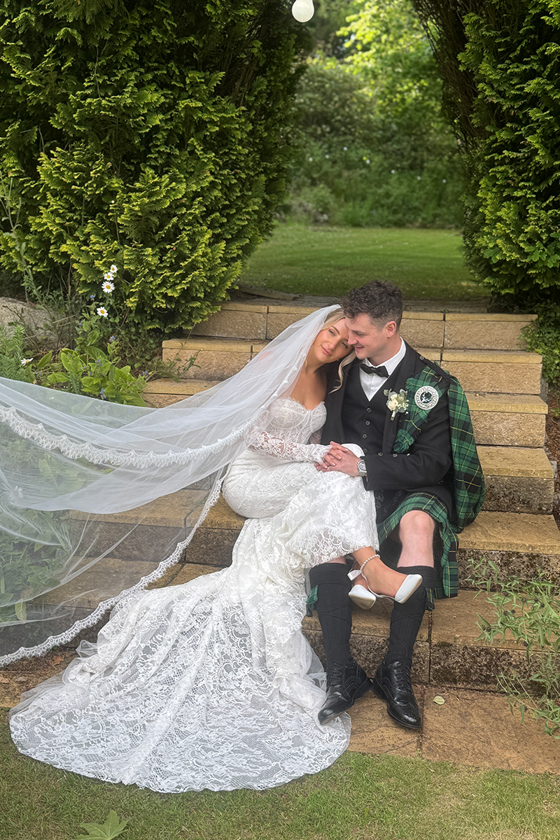 Bride rests her head on grooms shoulder while sitting on steps in gardens of wedding venue, captured by Scottish wedding content creator The Perfect Day Club.