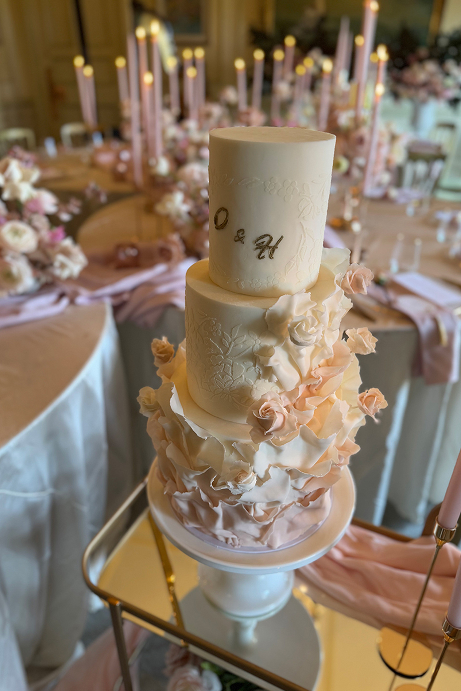 Cream three tier wedding cake with "O & H" in gold on its top tier, decorated with baby pink roses and flowers down to the bottom tier.