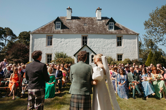 Bride and groom from behind during outdoor ceremony, with smiling guests and blue skies in background.
