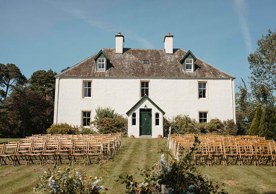 View of outdoor wedding with wooden chairs and a white house in background.