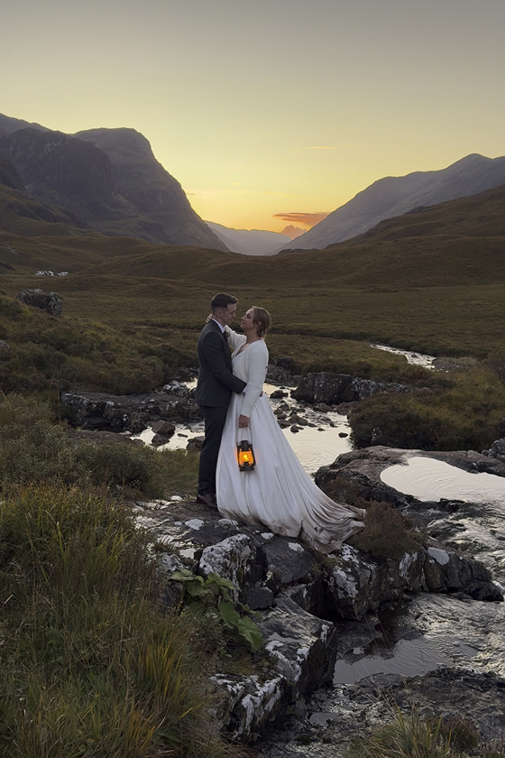 Newlyweds pose as bride holds a lantern glowing orange, with an orange sunset in the background behind the Scottish hills.