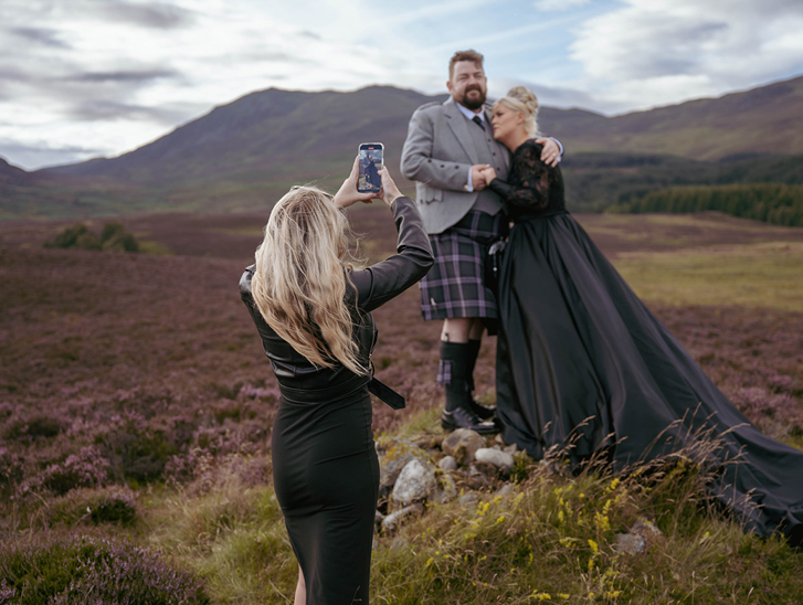 Louise, owner of Millar Moments, in action, capturing a dramatic image of a bride in a black wedding dress and her groom with Scottish hills as their backdrop.