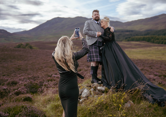 Louise, owner of Millar Moments, in action, capturing a dramatic image of a bride in a black wedding dress and her groom with Scottish hills as their backdrop.
