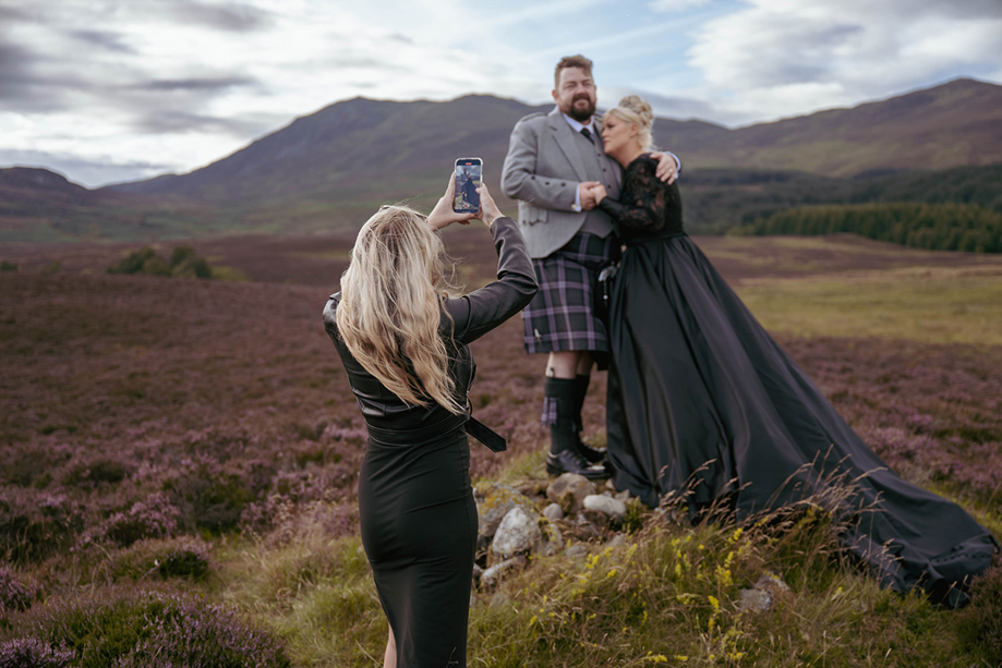 Louise, owner of Millar Moments, in action, capturing a dramatic image of a bride in a black wedding dress and her groom with Scottish hills as their backdrop.