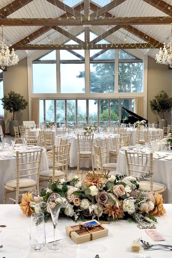 Image displaying wedding breakfast set-up with brown beams, orange and pink flowers and cream chiavari chairs.