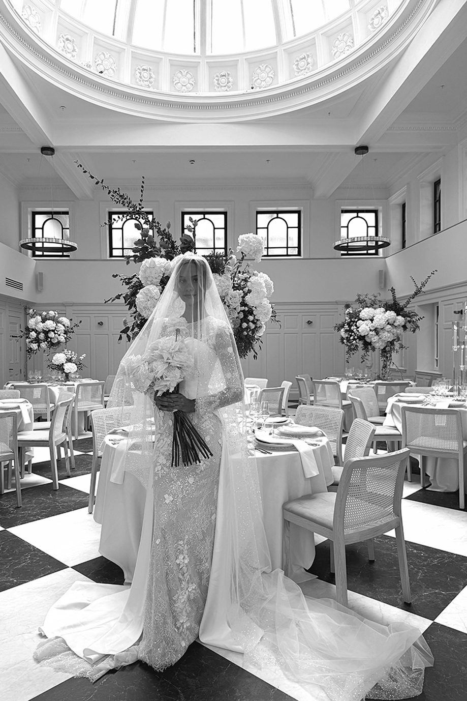 Black and white image of Emily Shak posing beneath The Collector's Hall's skylight with veil over her face and holding large bouquet, with wedding meal set-up in background.