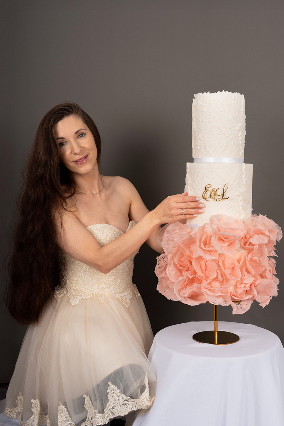 Owner of Eclectic Luxuries, Gabby, posing beside a three-tier cake with intricate detailing and pink chiffon base tier inspired by the bride's wedding dress.
