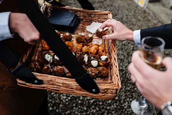 Tatties & Tipples team member holds wicker basket filled with canapés as guest holding glass of Champagne reaches for one.