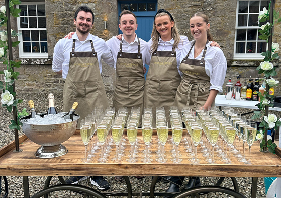 Four members of the Tatties & Tipples Event Catering team, standing in front of a cart filled with glasses of Champagne.
