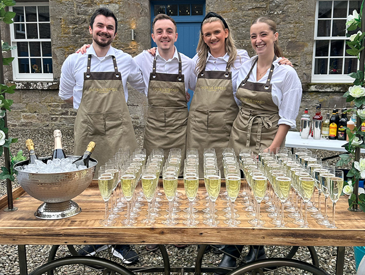 Four members of the Tatties & Tipples Event Catering team, standing in front of a cart filled with glasses of Champagne.