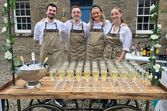 Four members of the Tatties & Tipples Event Catering team, standing in front of a cart filled with glasses of Champagne.