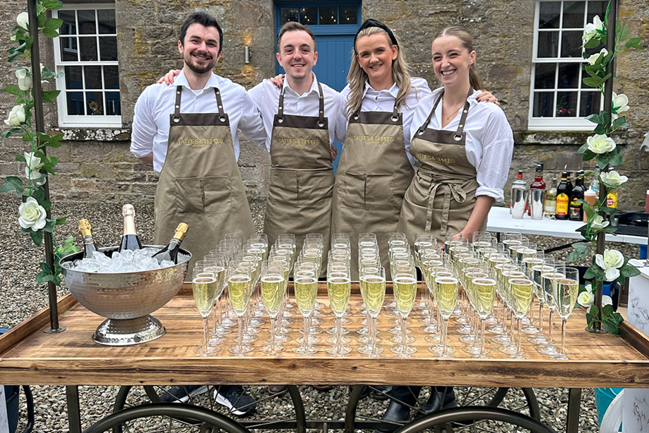 Four members of the Tatties & Tipples Event Catering team, standing in front of a cart filled with glasses of Champagne.