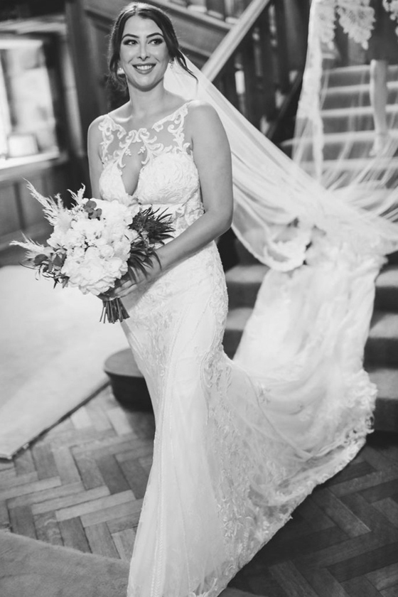 Bride smiling as she walks down a staircase wearing a lace wedding dress and veil, showcasing natural bridal makeup by Hannah Inglis Beauty