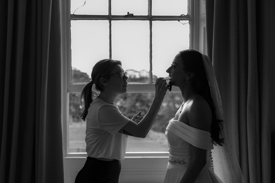 Bridal makeup artist Hannah Inglis applying makeup to a bride in front of a window during wedding morning preparations