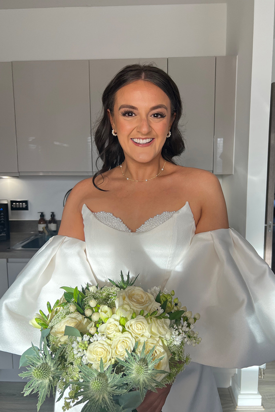 Bride wearing an off-the-shoulder puff-sleeve wedding dress holding a bouquet, with fresh bridal makeup by Hannah Inglis Beauty