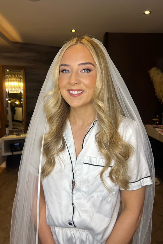 Bride with long blonde hair smiling in bridal pyjamas and veil during wedding morning makeup prep