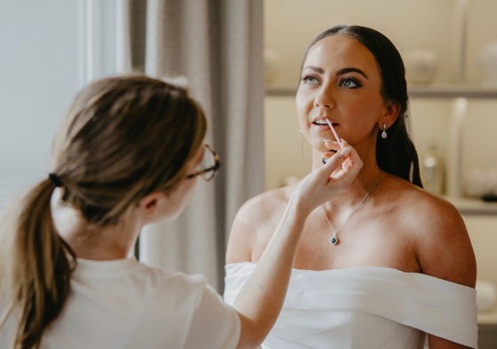 Wedding morning moment as Hannah Inglis applies lip gloss to a bride wearing an off-the-shoulder wedding dress