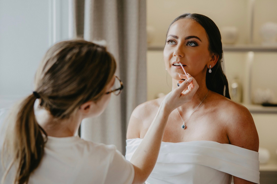 Wedding morning moment as Hannah Inglis applies lip gloss to a bride wearing an off-the-shoulder wedding dress