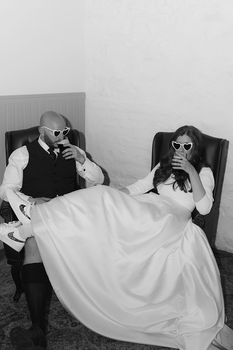 Bride and groom relax in leather armchairs wearing heart sunglasses while drinking Guinness during their Scottish wedding reception