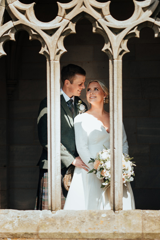 Newlyweds smiling at each other beneath carved stone arches during romantic Scottish castle wedding portraits